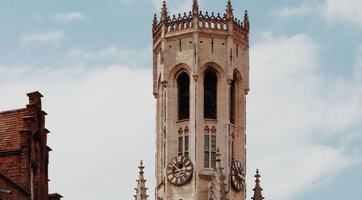 The medieval Belfry of Bruges rises above the city’s red-tiled rooftops, its clock faces framed against a partly cloudy blue sky.