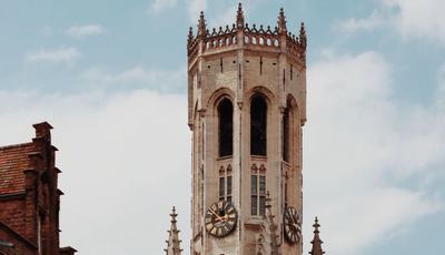 The medieval Belfry of Bruges rises above the city’s red-tiled rooftops, its clock faces framed against a partly cloudy blue sky.