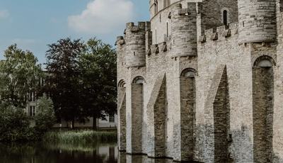Gravensteen Castle in Ghent rises beside its calm moat, with its medieval stone walls and round towers reflected in the water beneath a blue, partly cloudy sky.