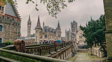 People cross Ghent’s historic Sint-Michielsbrug, with the ornate former Old Post Office and its clock tower on the left, Saint Nicholas’ Church in the centre and the Belfry rising behind the trees to the right.